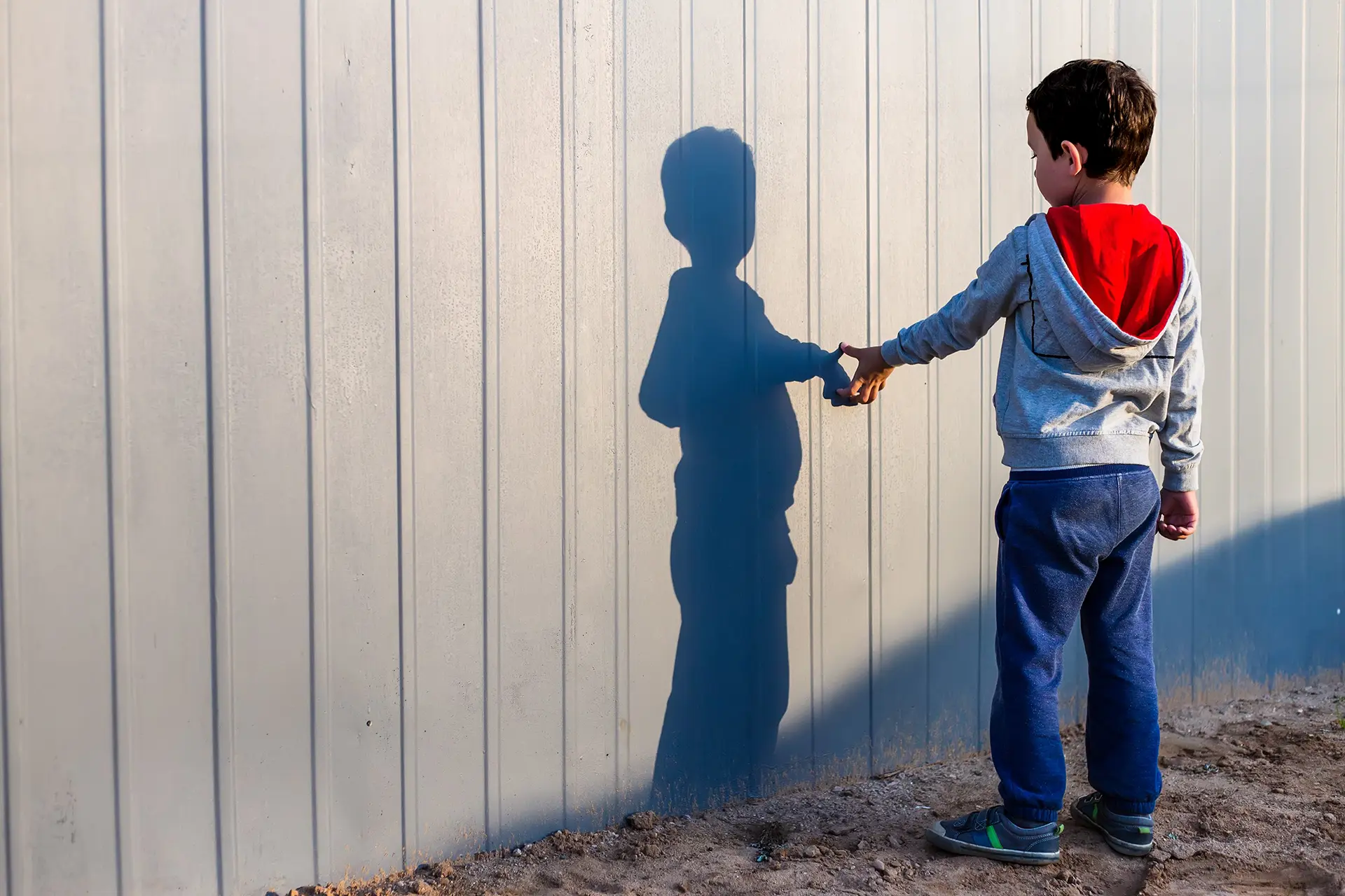 An autistic child standing outdoors near a wall, intently observing their own shadow reflected on the surface. The child is reaching out to touch hands with their shadow, attempting to shape their hands and shadow into a heart symbol, showcasing a moment of curiosity and self-expression.